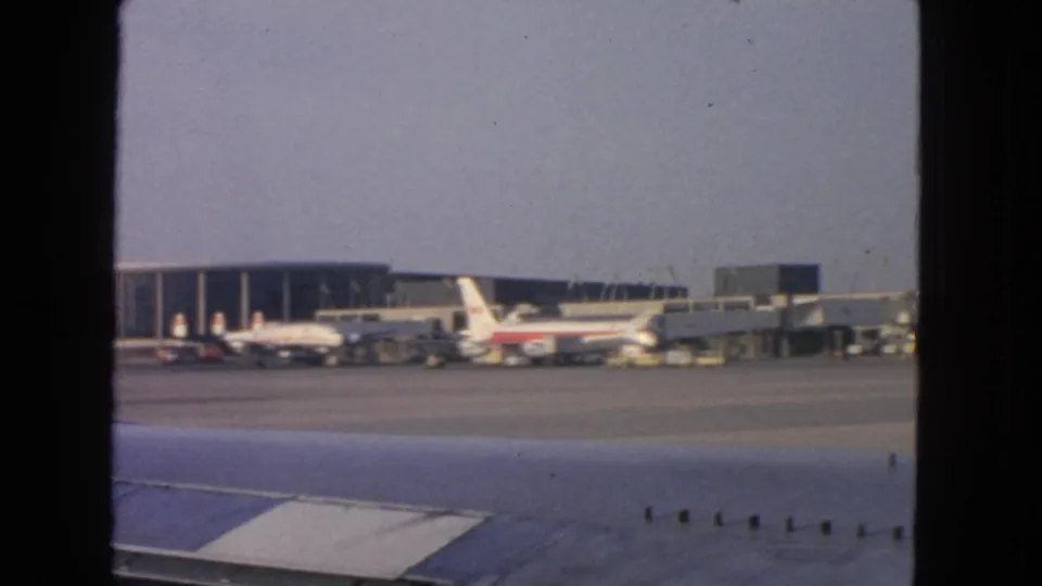 1963 San Francisco airport scene from a passenger window, showing the wing, parked jetliners, terminal gates, and tarmac activity.