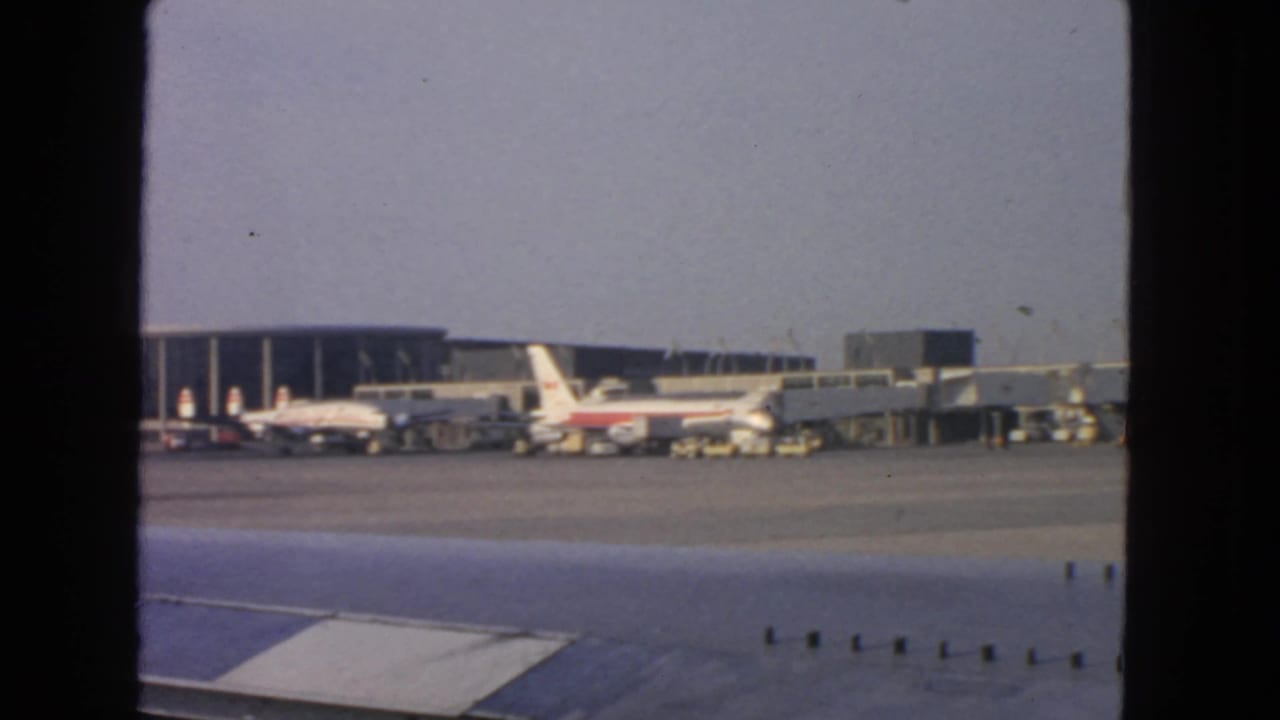 1963 San Francisco airport tarmac viewed from a passenger window, with an airplane wing in front and airliners at terminal gates.