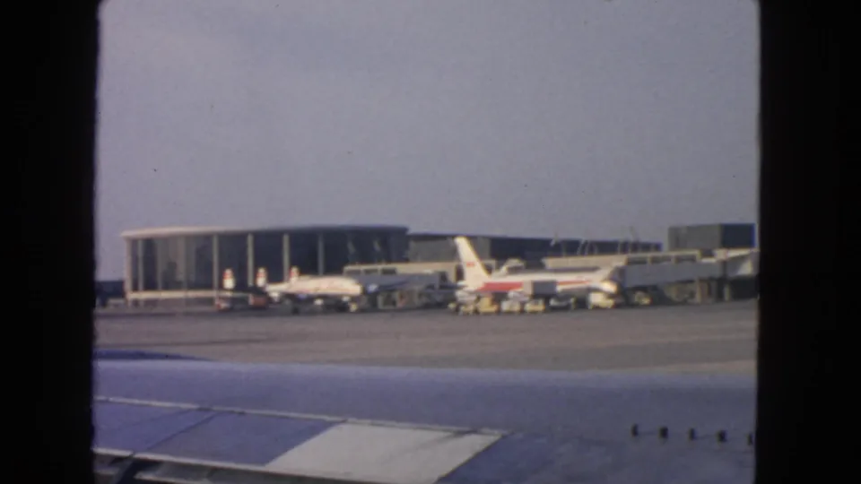 Soft 8mm view from an aircraft window toward a 1963 San Francisco airport terminal with parked airliners and ground equipment.