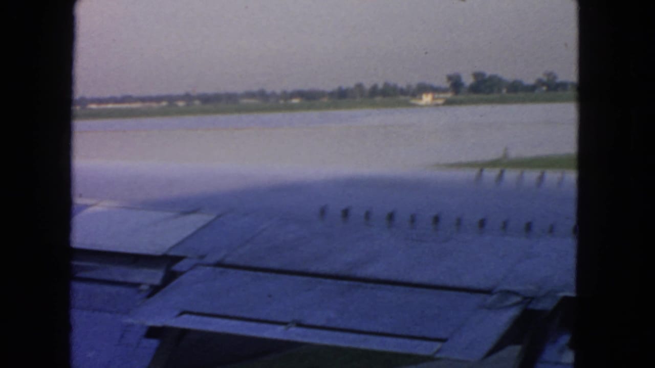 1963 San Francisco-area passenger-window view over an airplane wing, with water or runway edge and distant trees visible.