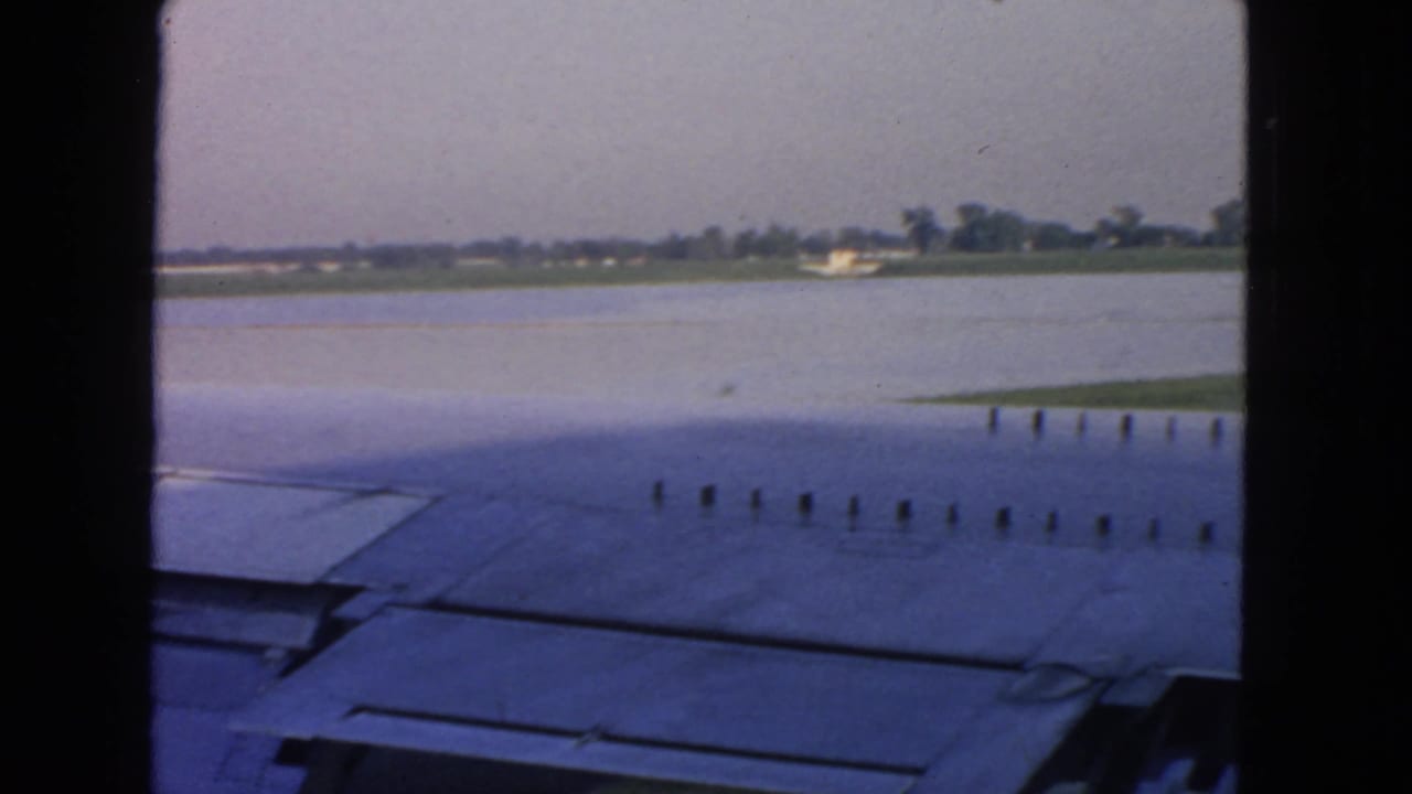 Aircraft wing and flap area fill the lower frame, with water and distant low buildings beyond.
