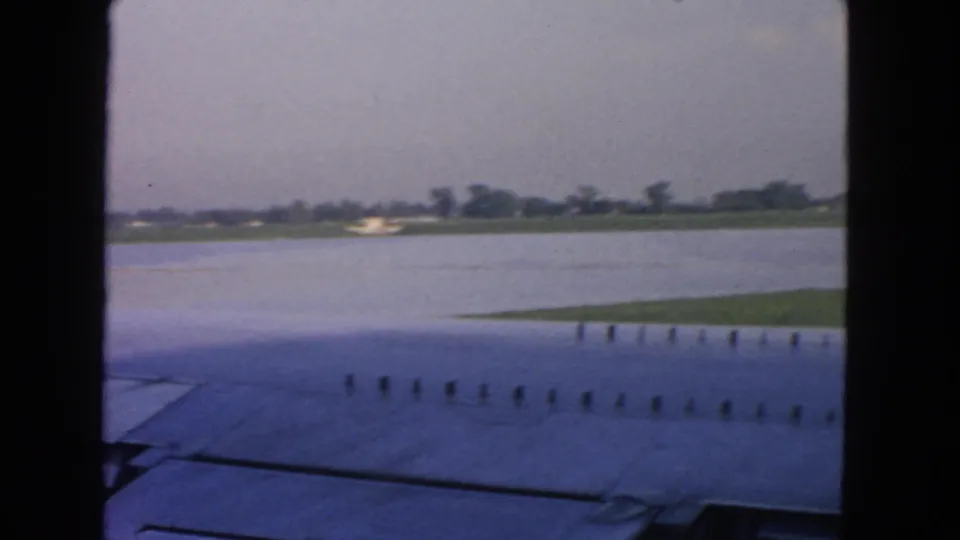 1963 home-movie view from an airplane window near San Francisco, looking past the wing toward water, grass, and distant airport structures.