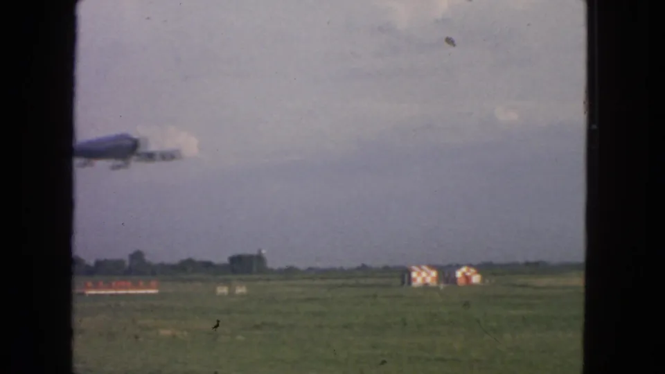 A plane in flight over a grassy San Francisco-area airfield, with low airport buildings and hazy sky visible in this 1963 footage.
