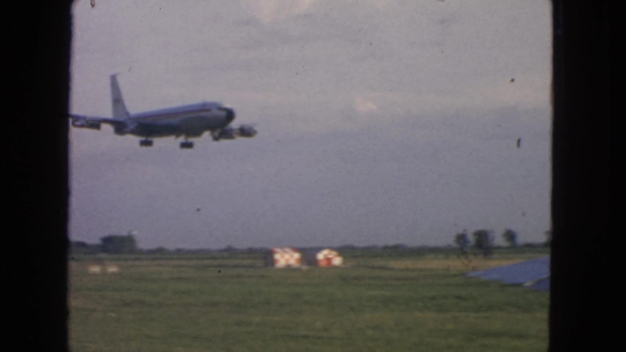 1963 8mm travel frame showing a jetliner descending over a grassy airfield, seen through dark window framing.