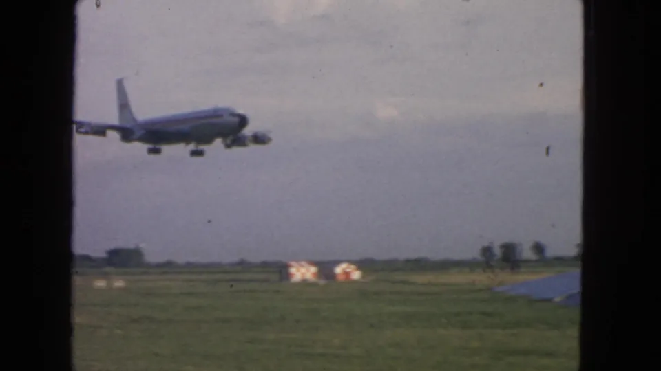 1963 8mm travel frame showing a jetliner descending over a grassy airfield, seen through dark window framing.
