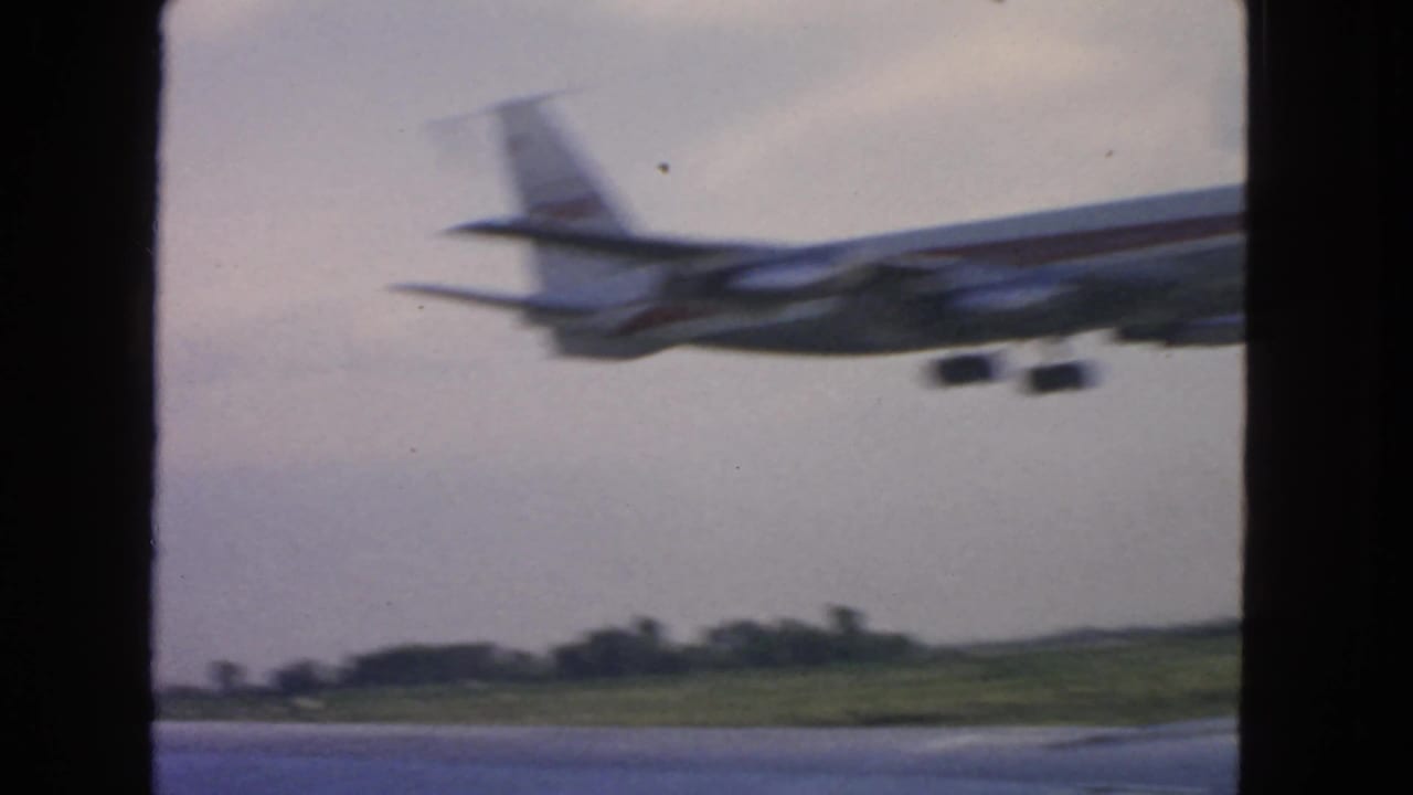 Blurred jetliner passing low over runway-side grass and distant trees.