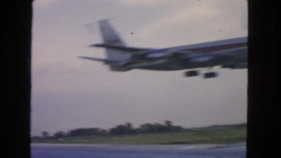 1963 8mm frame of a jetliner passing close to the runway, with motion blur and a rural airfield background.