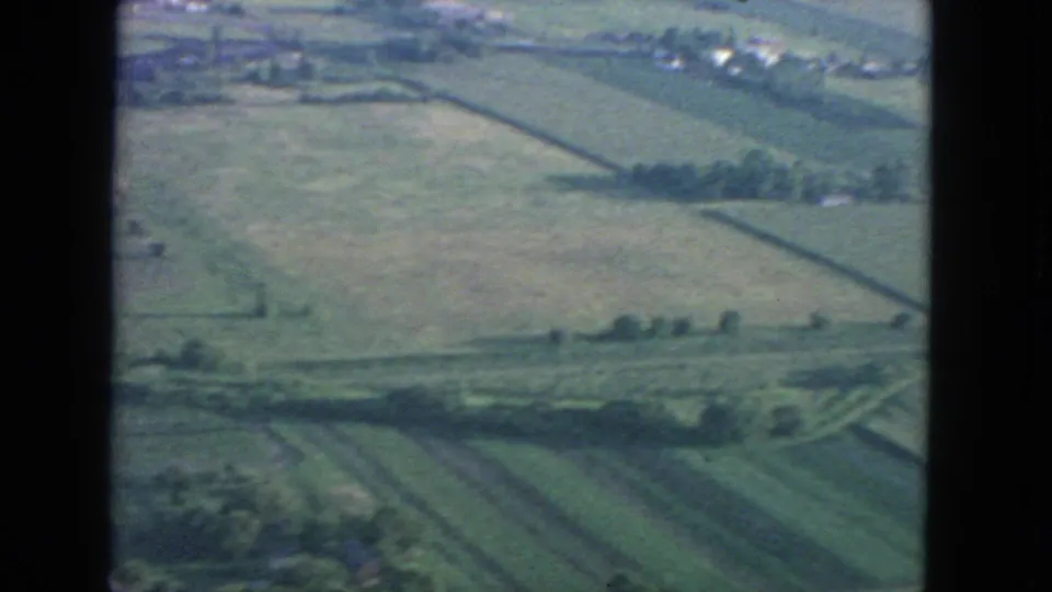 1963 8mm aerial frame showing patchwork farmland, field borders, and distant buildings from above.