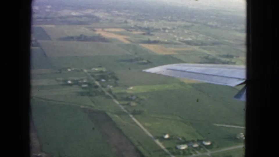 1963 home-movie aerial view with a wing tip at right, showing farmland, narrow roads, and small rural structures below.