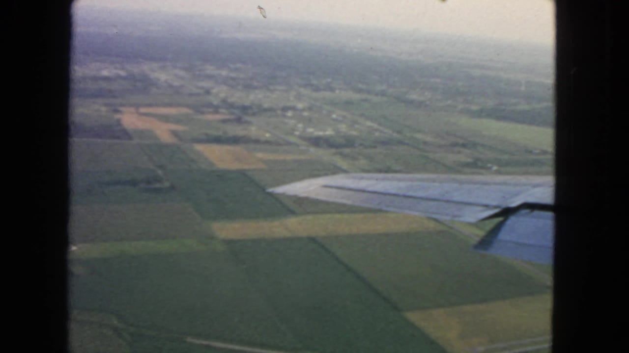 Airplane wing over green and brown fields with distant houses under a hazy sky.