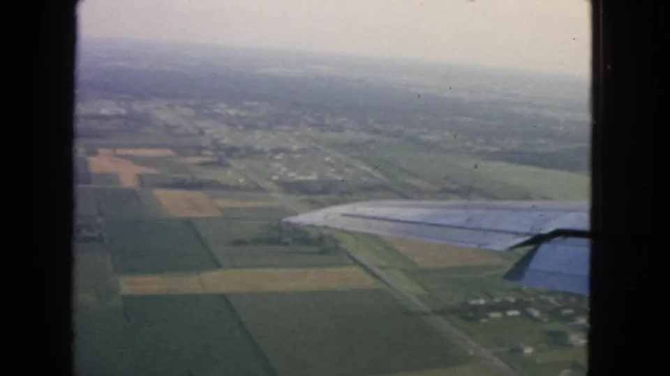 Airplane-window view over patchwork farmland and a small developed area, with the wing visible at right in 1963 home-movie footage.