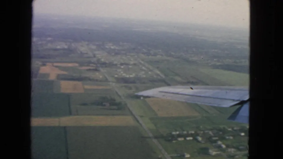 1963 8mm aerial view from a passenger plane, showing fields, roads, scattered homes, and the aircraft wing near the lower edge.