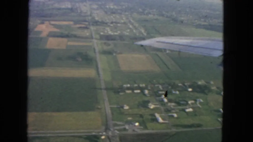A passenger-filmed 8mm aerial view over farmland, roadways, and scattered buildings, with the airplane wing crossing the upper right.