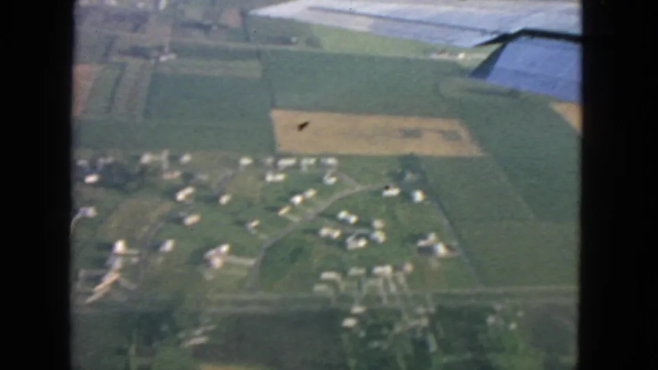Soft 8mm aerial view from a plane, with rural houses, green fields, and a wing edge in a 1963 San Francisco-area travel clip.