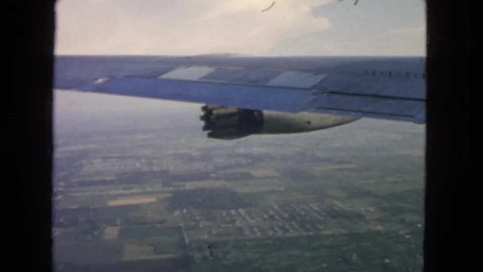 1963 8mm passenger view of an airplane wing and engine above distant farmland and haze near San Francisco.