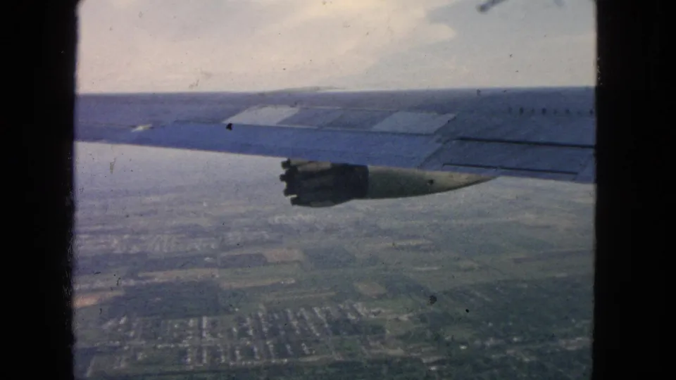 Vintage 8mm aircraft-window view with jet wing, engine, haze, fields, and suburban housing below near San Francisco.