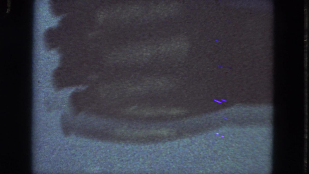 1963 San Francisco 8mm frame with a dark obscured industrial-looking form, visible film edge, grain, scratches, and blue-purple speckles.