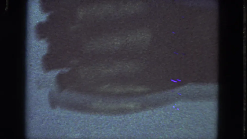 1963 San Francisco 8mm frame with a dark obscured industrial-looking form, visible film edge, grain, scratches, and blue-purple speckles.