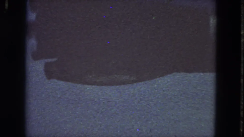 1963 San Francisco 8mm frame showing an indistinct dark horizontal form, light textured background, heavy grain, scratches, and colored aging speckles.