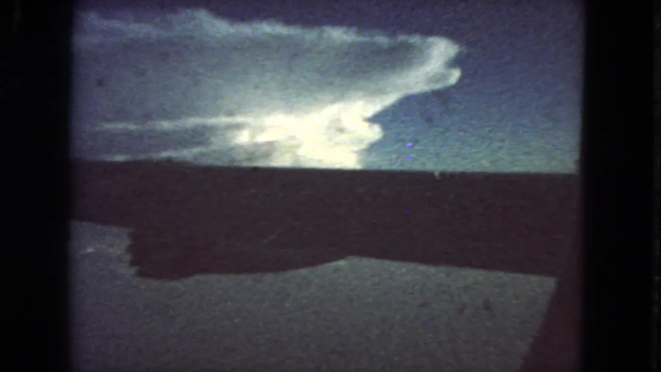 Passenger-window view from a 1963 8mm flight, showing the aircraft wing as a dark band against pale clouds and blue sky.