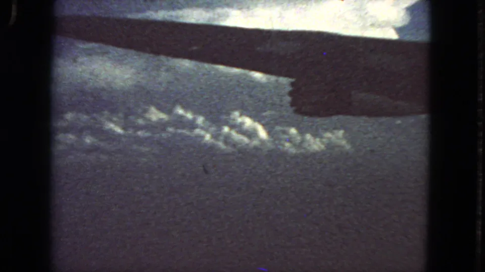 1963 home-movie frame from an airplane window, with the wing underside cutting across a textured cloud layer and open sky.