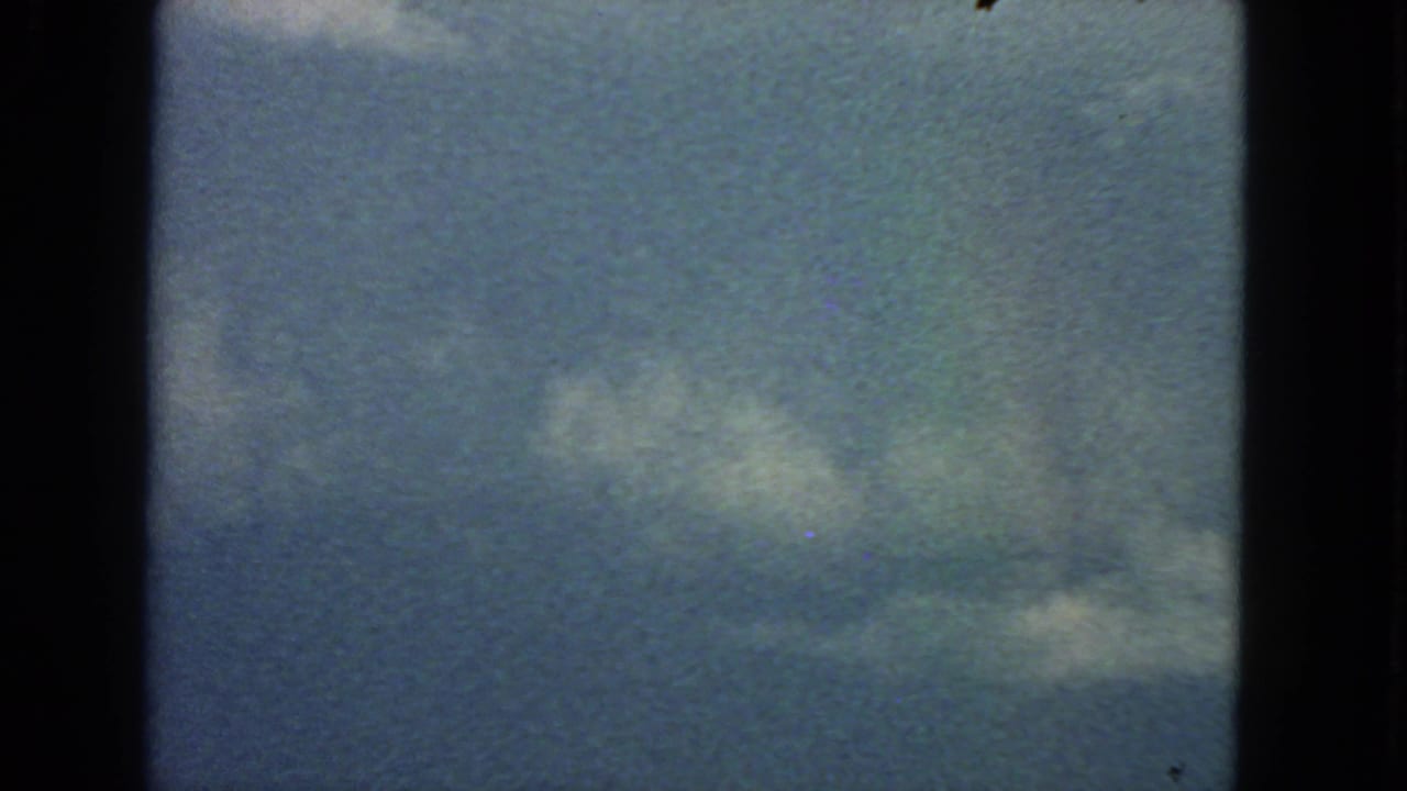 1963 San Francisco 8mm frame looking up at blue sky, scattered clouds, film grain, and a faint rainbow color trace.