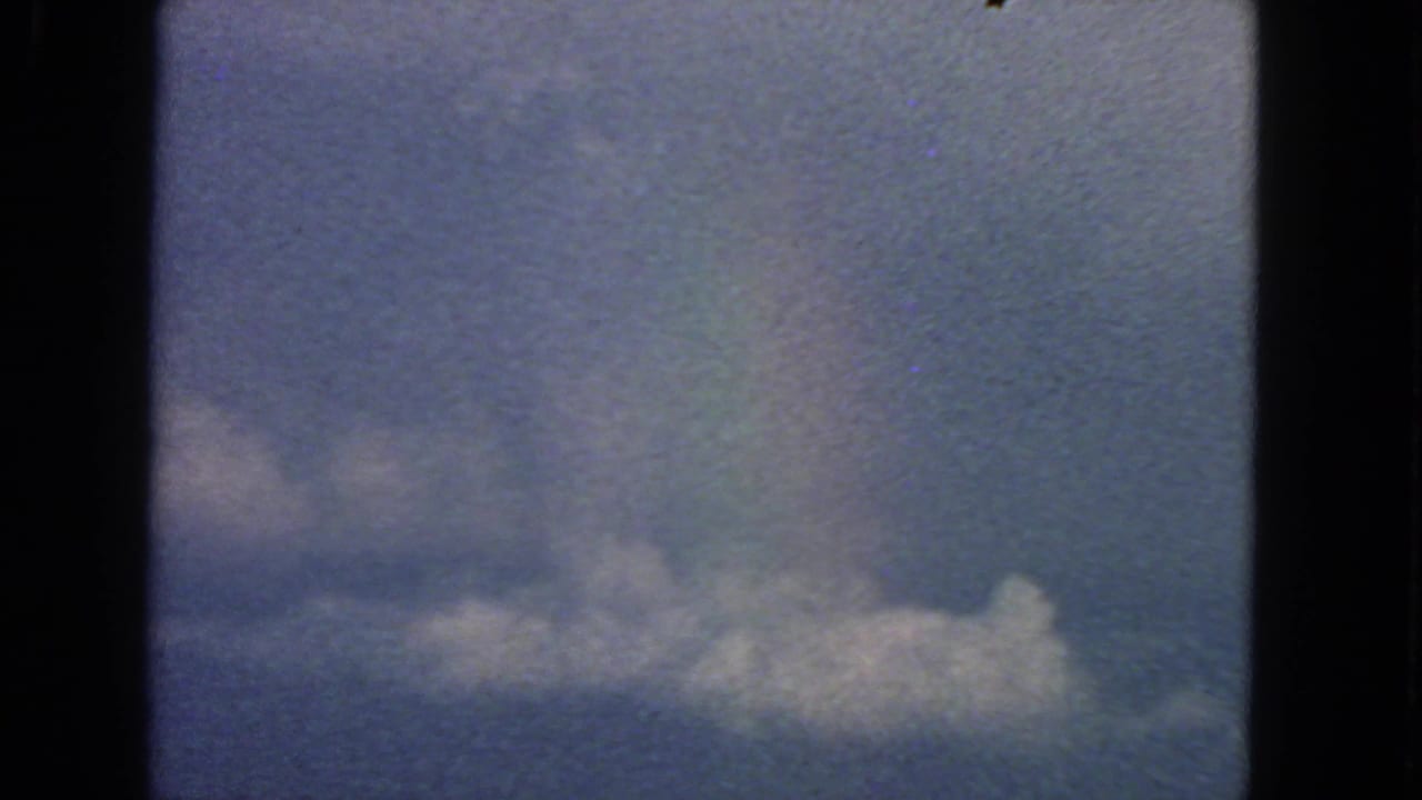 1963 8mm airplane-window view near San Francisco showing blue sky, a horizontal cloud bank, dark frame edges, and film grain.