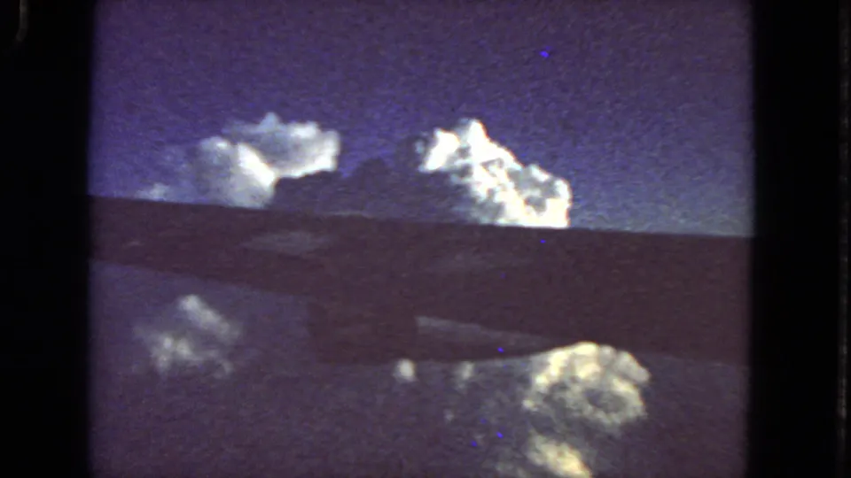 1963 8mm airplane-window perspective with clouds, reflected highlights, and a dark wing or window edge crossing the frame.