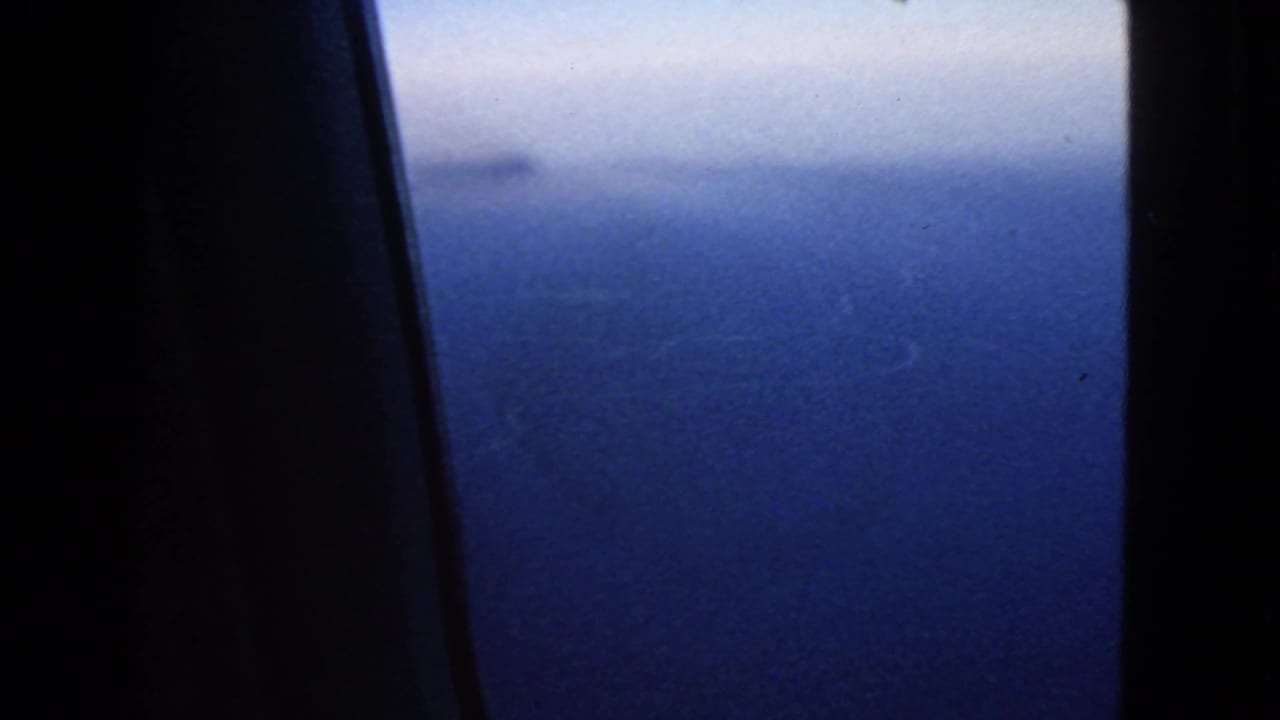 1963 8mm view of blue ocean and hazy horizon through an airplane window, with dark interior framing at the left.