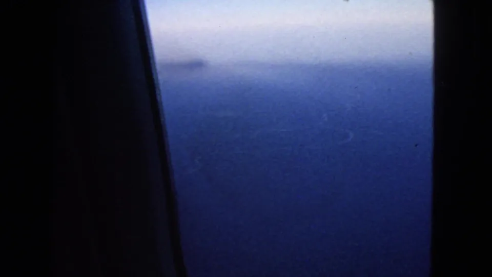 1963 8mm view of blue ocean and hazy horizon through an airplane window, with dark interior framing at the left.