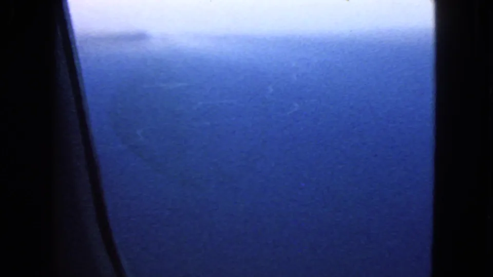Aerial ocean view from a 1963 home movie, with a dark airplane window support cutting through the blue water scene.