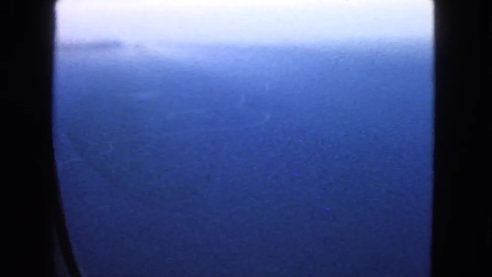 1963 8mm aerial view over blue water, partly obscured by dark airplane window edges and a soft hazy horizon.