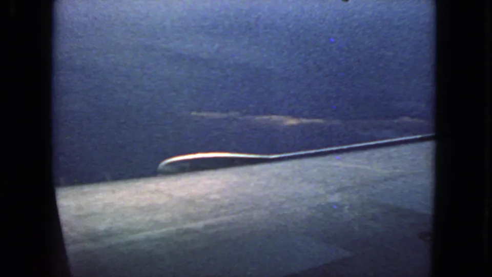 1963 8mm frame with dark airplane-window borders, blue sky, and a pale wing-like aircraft surface across the lower portion.