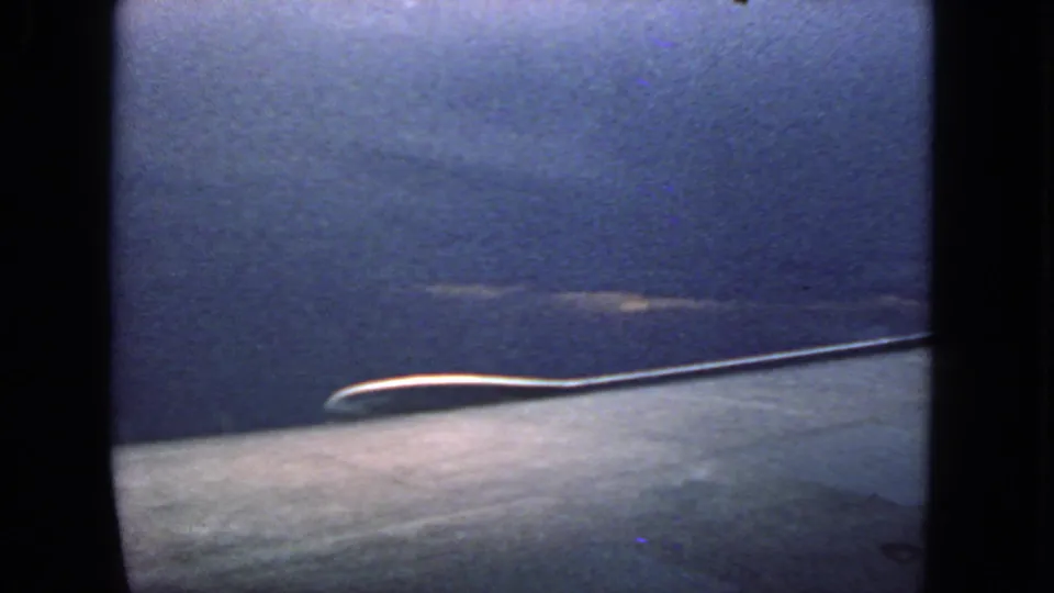1963 8mm passenger view toward blue sky, with a pale cloud band and the curved airplane window edge visible.