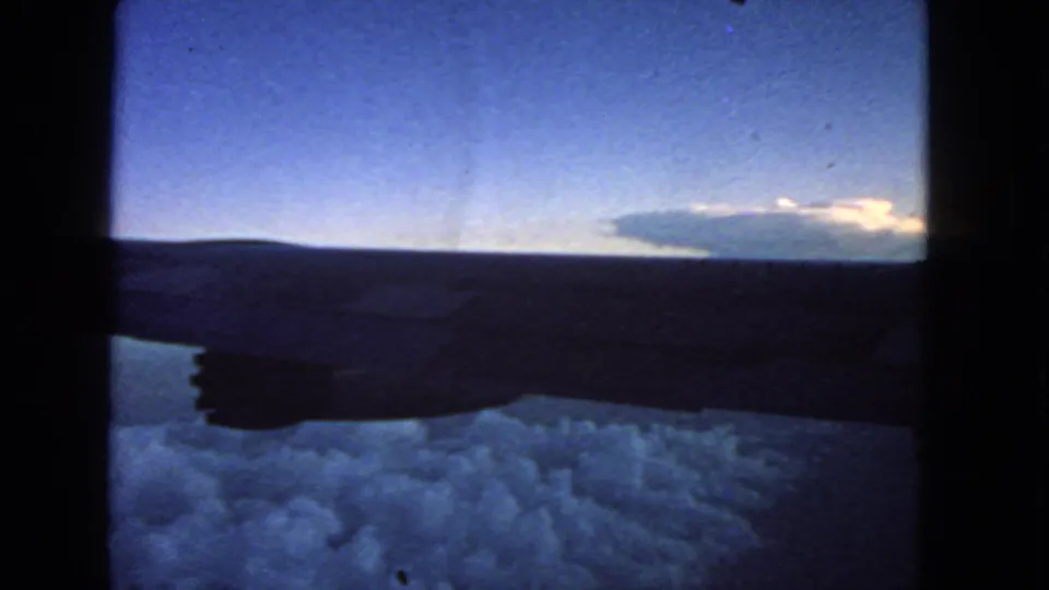 1960s home-movie frame from an airplane window, showing the wing silhouette, clouds below, and dusk light on the horizon.
