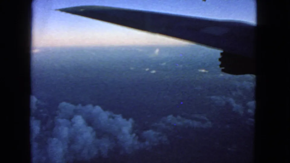 Passenger-window view of a dark airplane wingtip above clouds, with dusk sky and distant light in archival 8mm footage.