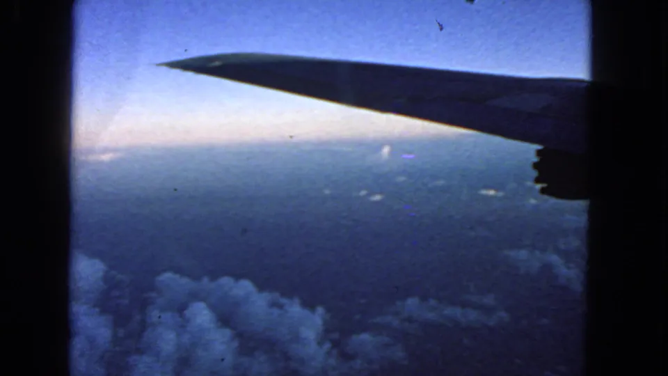 1963 8mm airplane-window view with a dark wing silhouette over clouds and a blue horizon, metadata placing the trip near San Francisco.
