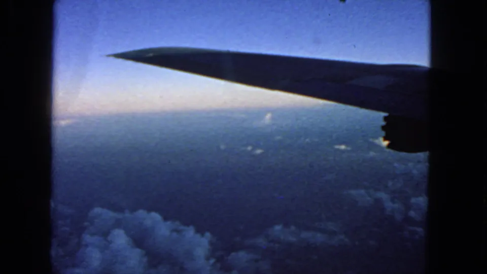 Airplane wing viewed from a passenger window over cloud cover, with 8mm grain and blue color shift from the 1963 home movie.