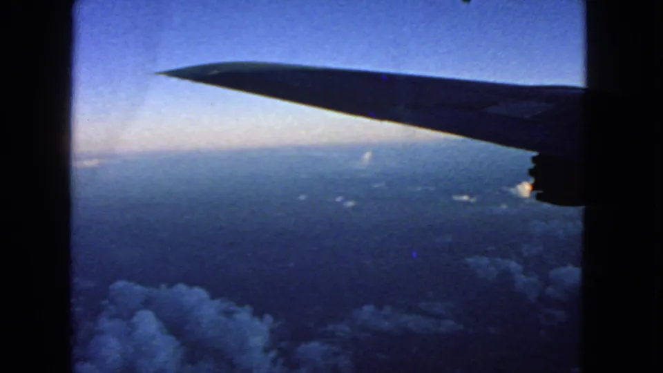 Vintage 8mm frame of an airplane wing over a hazy horizon and cloud tops during the 1963 flight footage.