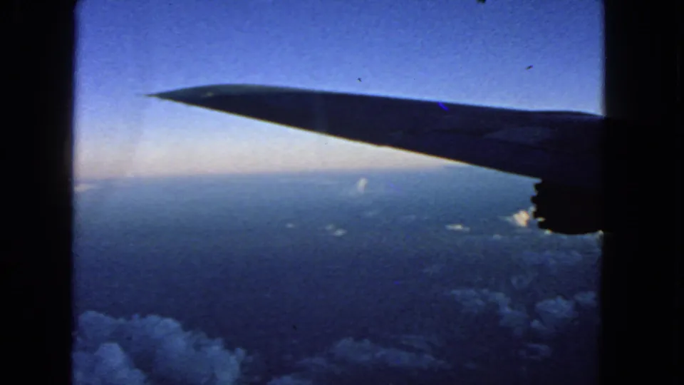 Aircraft wing and partial underside detail silhouetted against blue sky above clouds, from the 1963 8mm airplane-window scene.