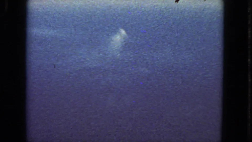 Blue-tinted 8mm aerial frame with faint cloud shapes and heavy film grain during the 1963 passenger flight.