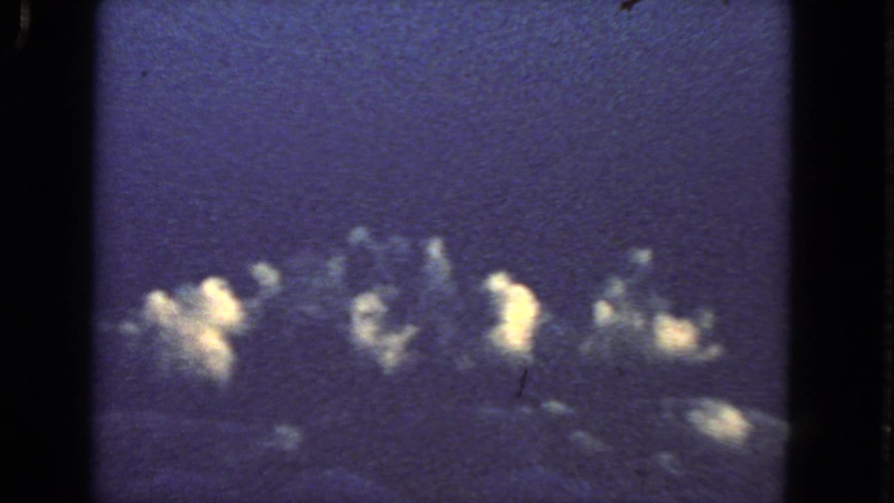 Dark airplane window framing with blue sky, clouds, and bright irregular flashes near the horizon.