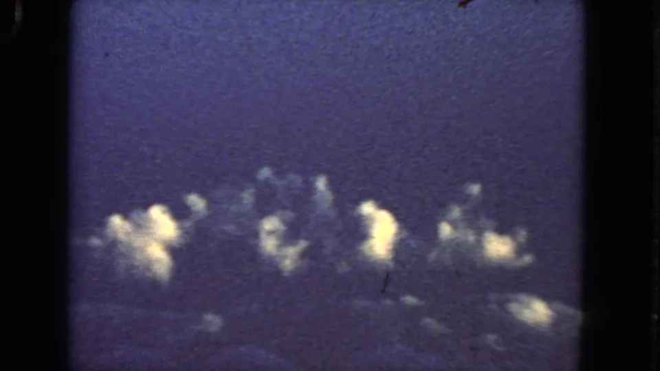 1963 8mm airplane-window view near San Francisco metadata, showing blue sky, cloud tops, and bright uncertain flashes beyond the window frame.
