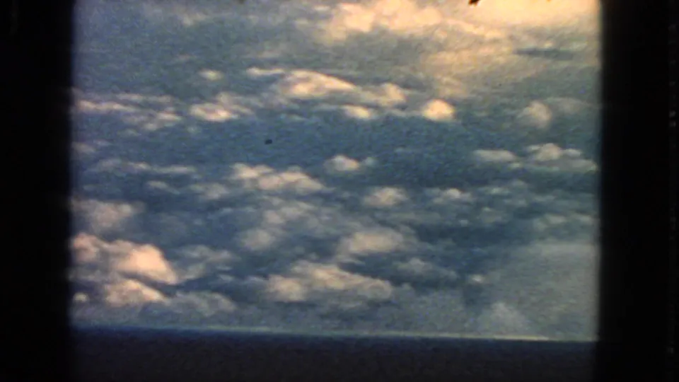 1963 8mm aerial view showing sunlit cloud tops and blue atmosphere through a dark aircraft window, with no people or landmarks visible.