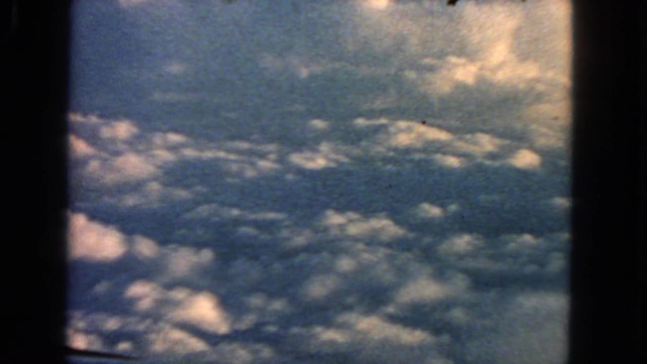 1963 8mm home-movie view from an airplane window over sunlit cumulus clouds, with dark frame edges visible.