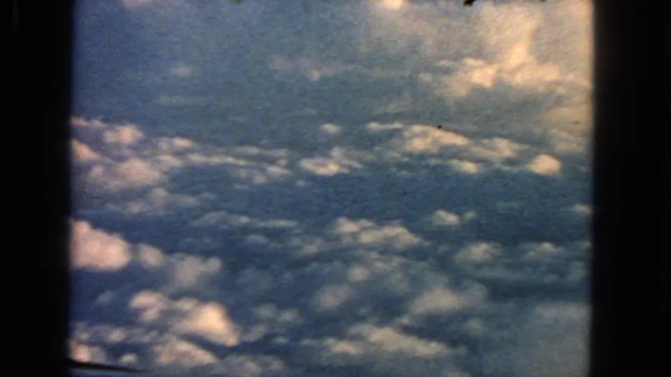 1963 8mm home-movie view from an airplane window over sunlit cumulus clouds, with dark frame edges visible.