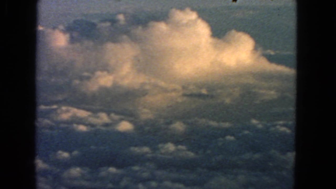 1963 8mm home-movie view of bright clouds and blue sky, with dark frame edges around the aerial scene.
