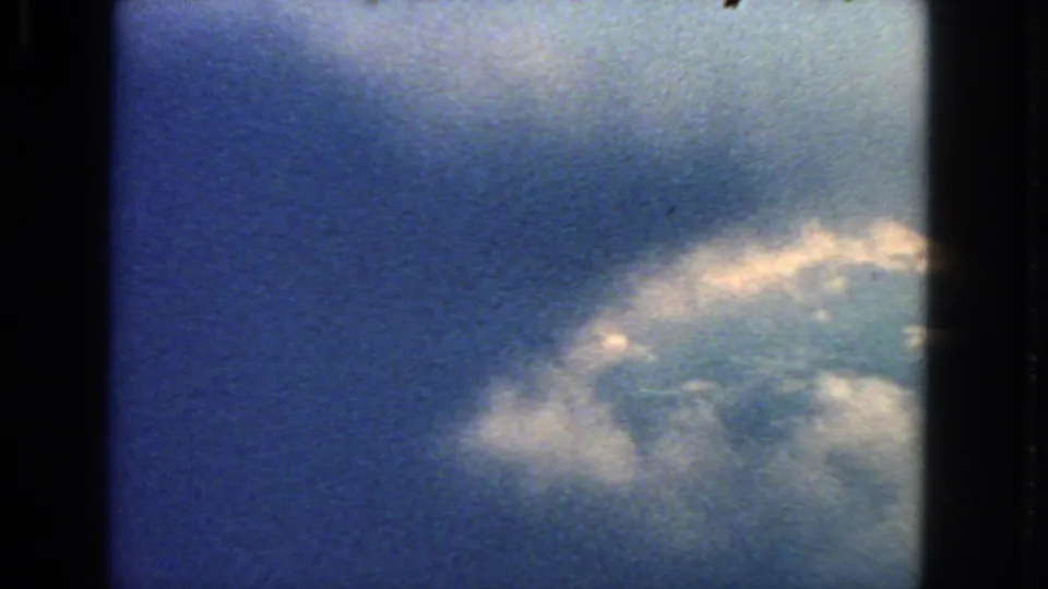 1963 home-movie aerial view with white cloud tops crossing the lower frame under a blue sky.