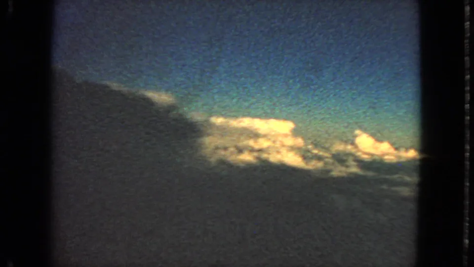 1963 8mm in-flight view of bright cloud banks and blue sky through a dark window edge.