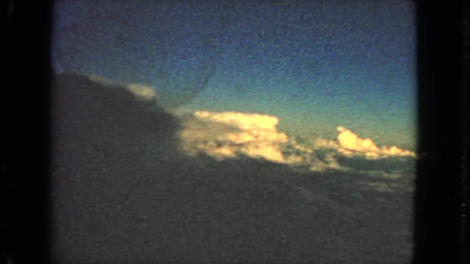 1963 8mm airplane-window view with a dark side frame, blue sky, and glowing clouds.